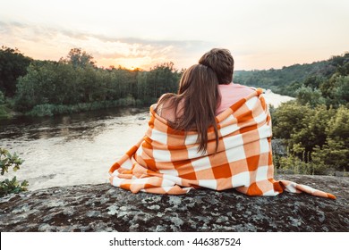 Loving couple wrapped in plaid sitting on mountain outdoors on sunset. Rear view. Man and woman traveling together. Traveler couple enjoying nature, embracing, watching landscape under a blanket. - Powered by Shutterstock