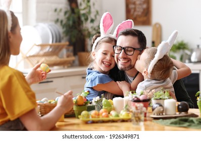 Loving cute children wearing funny bunny ears headbands embracing and kissing cheerful young father while gathering with family at table in kitchen for Easter eggs painting - Powered by Shutterstock