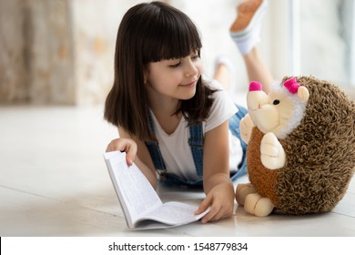 Loving Cute Little Girl Lying On Warm Home Floor Reading Interesting Story To Toy Hedgehog Friend, Happy Small Preschooler Kid Have Fun Playing Learning With Stuffed Animal, Enjoy Fairy Tale In Book