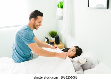 Loving dad smiling to a little girl and tucking her in bed at night. Adorable daughter telling goodnight to her father