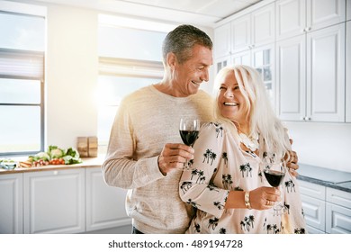 Loving elderly couple enjoying a happy retirement standing arm in arm smiling and laughing as they drink a glass of red wine together in the kitchen - Powered by Shutterstock