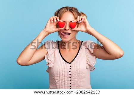 Loving eyes. Beautiful young adult woman with two funny hair buns holding two valentine hearts in front of her eyes like glasses, shows tongue out. Indoor studio shot isolated on blue background.