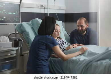 Loving Family Comforting Under Treatment Sleeping Ill Kid In Hospital Pediatric Ward. Considerate Parents Sitting Beside Hospitalized Sick Little Daughter Inside Children Healthcare Facility Room.