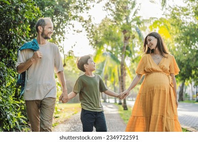 A loving family enjoying a leisurely walk in the park - a radiant pregnant woman after 40, embraced by her husband, and accompanied by their adult teenage son, savoring precious moments together - Shutterstock ID 2349722591
