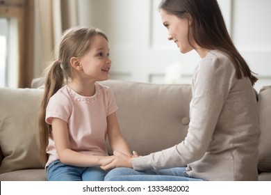 Loving family happy mother and cute child girl holding hands talking sitting on sofa at home, caring elder sister mom baby sitter having friendly trust conversation with preschool little kid daughter