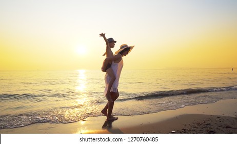 Loving Family Having Fun On The Beach. Boy Riding Piggy Ride On Mothers Back With Waves Splashing Her Feet Against Ocean Sunset. Steadicam Shot