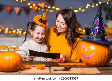 loving family  mom   daughter  cut out paper figures  making decorations for house for Halloween  at wooden table in kitchen  woman   girl draw bats   pumpkins  lights garlands in background