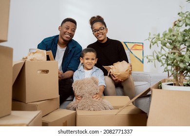 A Loving Family Sits On The Couch In Living Room And Unpacks Boxes After Moving To A New Apartment, They Pull Out Old Memorabilia, Paintings, Parents And Son Smile At The Camera