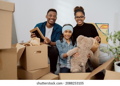 Loving Family Sits On Couch In Living Room Unpacks Multitude Of Boxes Standing Around Them After Moving To New Apartment They Pull Out Old Memorabilia Parents With Their Daughter Smile At Camera