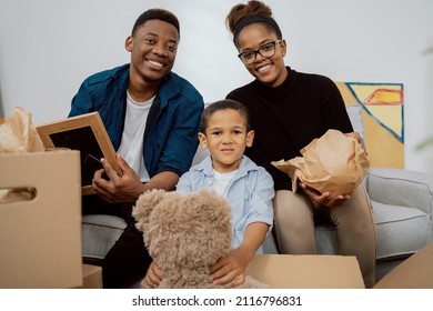 A Loving Family Sits On The Couch In Living Room And Unpacks Boxes After Moving To A New Apartment, They Pull Out Old Memorabilia, Paintings, Parents And Son Smile At The Camera