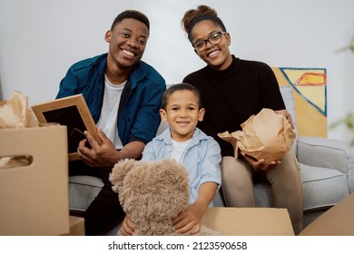 A Loving Family Sits On The Couch In Living Room And Unpacks Boxes After Moving To A New Apartment, They Pull Out Old Memorabilia, Paintings, Parents And Son Smile At The Camera