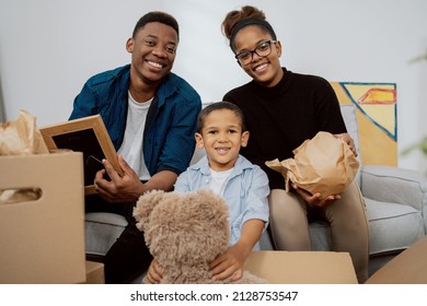 A Loving Family Sits On The Couch In Living Room And Unpacks Boxes After Moving To A New Apartment, They Pull Out Old Memorabilia, Paintings, Parents And Son Smile At The Camera