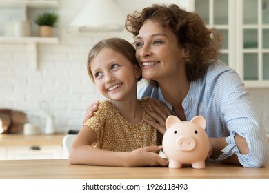 Loving Family Of Young Mother Little Daughter Hug By Kitchen Table Hold Cute Piggybank Look Away Plan Happy Future Life. Dreamy Mom Small Child Saving Money For Future Vacation Getting Good Education