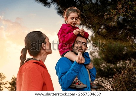 A loving father carries his daughter on his shoulders and shows her a freshly picked wild berry, teaching her about nature during a family hiking adventure at sunset.