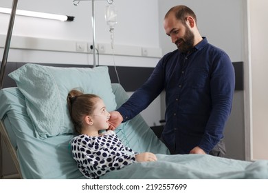 Loving Father Comforting Hospitalized Sick Daughter Resting In Patient Bed Inside Hospital Pediatric Ward Room. Caring Parent Taking Care Of Ill Little Girl While Touching Her Face.