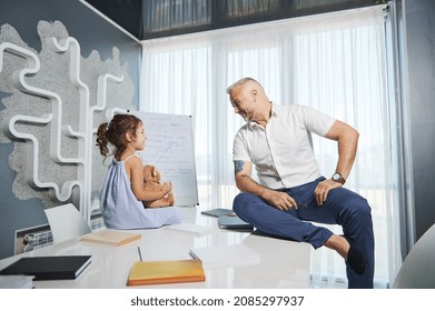 A Loving Father Communicates With His Cute Little Daughter Sitting On The Table. Concept Of Love, Care And Parental Attention Regardless Of Busy Work Schedule