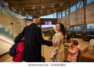 Loving Father Hugs His Son And Wife As She Smiles, Looking Over Her Shoulder, Pointing To Timetable Information Panel, Checking The Flight At The Airport Departure Terminal. Family Air Travel Concept