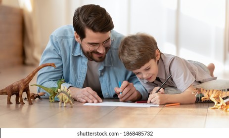 Loving father lying on floor at home drawing painting with small son enjoy family weekend together. Caring dad have fun play rest engaged in funny activity with little boy child in living room.