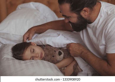 Loving father tucking blanket for his lovely little daughter. Adorable girl sleeping in bed, hugging her teddy bear, her father pulling up her blanket. Single parenting concept - Powered by Shutterstock