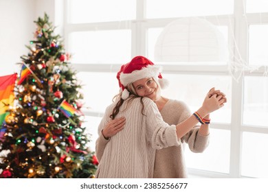 Loving female couple dancing at home interior. Two women in Santa Claus hats embracing at living room by rainbow flag and Christmas tree. LGBT family concept - Powered by Shutterstock