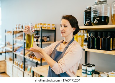 Loving female shop assistant in apron holding glass bottle with olive oil over shelves with eco packaging free products working in a zero-waste shop. No plastic conscious minimalism vegan lifestyle - Powered by Shutterstock