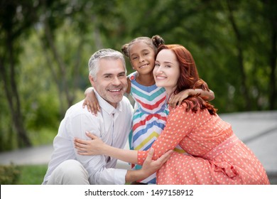 Loving foster parents. Loving foster parents feeling cheerful while hugging their lovely dark-skinned girl