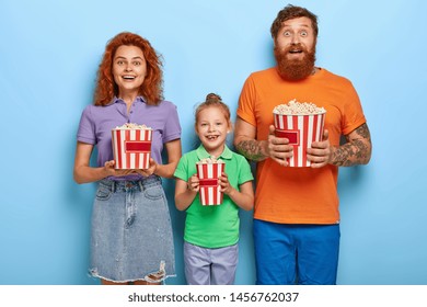 Loving Ginger Family Of Three Members Eat Popcorn While Watch TV Programme, Look Happily At Camera. Mother, Father And Daughter Spend Leisure Time At Local Cinema, Enjoy Watching Funny Comedy