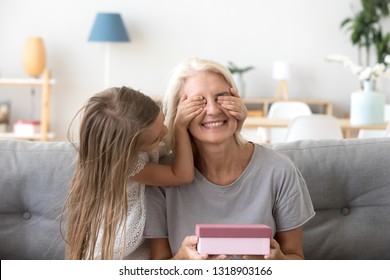 Loving Granddaughter Making Surprise To Happy Grandmother On Birthday Or Mothers Day, Preschool Girl Closing Eyes Of Excited Grandma Receiving Present, Holding Box With Gift, Sitting On Sofa At Home