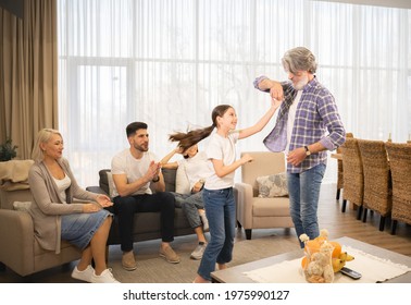 Loving Grandfather Dancing With Cute Little Girl Granddaughter In Front Of Grandmother And Parents Relaxing On Sofa In Living Room At Home. Happy Family Enjoying Leisure Time And Having Fun Together