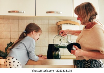 Loving Grandmother Cooking Together With Granddaughter At Home, Using Blender To Mix Vegetables, Little Girl Learning How To Use Appliances With Grandma In White Kitchen Environment. Family Concept