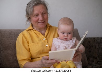 Loving Grandmother Reading A Book To Baby Girl Holding Book Sitting On Sofa. Nanny Granny Telling Story To A Grandchild