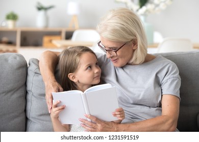 Loving Grandmother Teaching Granddaughter Holding Book Sitting On Sofa, Grandma Baby Sitter Embracing Kid Girl Reading Fairytale To Cute Child, Nanny Granny Telling Story To Preschool Grandchild