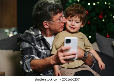 Loving Grandpa Sitting On A Sofa, Trying To Take Selfie With His Toddler Grandson. Boy Stuck His Tongue Out. Christmas Tree In Background.