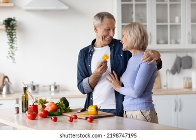 Loving Handsome Senior Man Husband Feeding His Smiling Pretty Wife While Cooking Delicious Healthy Meal, Happy Elderly Spouses Embracing, White Cozy Kitchen Interior, Copy Space