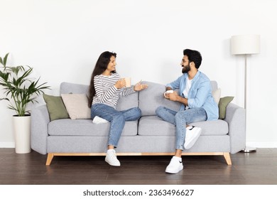 Loving happy beautiful young eastern couple sitting on couch in cozy living-room, drinking coffee and have conversation, cheerful man and woman enjoying weekend together at home, full length - Powered by Shutterstock