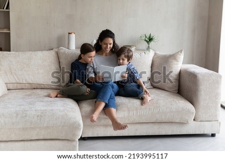 Loving happy caring young mother cuddling little cute kids son daughter, reading fairy tale stories in paper book. Happy two generations family enjoying leisure domestic activity, childcare concept.