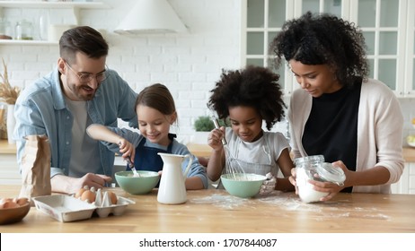 Loving Happy Diverse Parents With Little Multiracial Daughters Baking In Modern Kitchen, Smiling Multiethnic Young Family With Small Girls Children Cook Together Prepare Pancakes Or Biscuits At Home