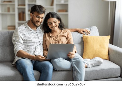 Loving happy indian couple sitting on couch at home, embracing, using laptop computer, young man and woman watching movie online, copy space - Powered by Shutterstock