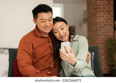 Loving happy korean spouses video calling via cellphone, middle aged man and his young wife relaxing on sofa at home, browsing new app or shopping online - Powered by Shutterstock