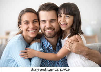Loving happy man embracing wife and little child daughter looking at camera, smiling mom dad hugging cute kid girl posing together on fathers day, cheerful young family of three headshot portrait