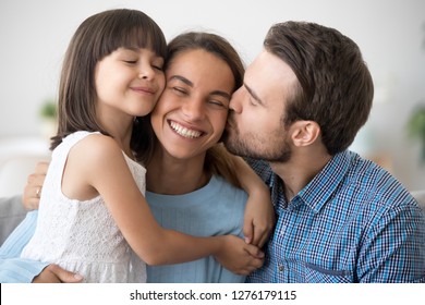 Loving Husband And Cute Kid Daughter Embracing Kissing Happy Mom Wife On Cheek Congratulating With Mothers Day, Little Child Girl And Dad Hugging Smiling Mum, Caring Family Of Three Bonding Together