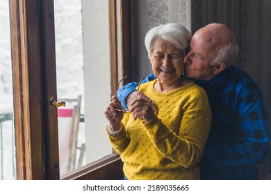 Loving husband hugging his gray-haired overjoyed wife from behind and softly kissing her - Sweet senior couple standing near the window and enjoying winter season together. High-quality photo - Powered by Shutterstock