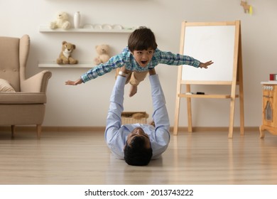 Loving Indian father lifting excited little son pretending flying, lying on warm wooden floor at home, happy carefree dad and preschool kid having fun together, involved in funny active game
