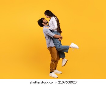 Loving Indian Guy Lifting His Girlfriend And Holding Her On Yellow Studio Background, Profile Side View, Free Space Banner. Romantic Couple Expressing Their Love To Each Other