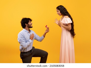Loving indian man standing on one knee and offering engagement ring to his beloved woman on yellow studio background. Young guy making proposal to sweetheart on Valentine's Day