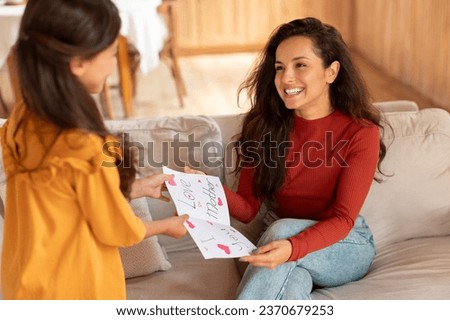 Loving kid daughter greeting mom with Mothers Day giving handcrafted card as a present, spending time with mommy and celebrating holiday together at modern home interior. Selective focus