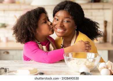 Loving Little Daughter Kissing Her Happy African American Mom In Kitchen, Cheerful Black Family In Aprons Posing While Baking At Home Together, Having Fun And Enjoying Homemade Pastry, Closeup