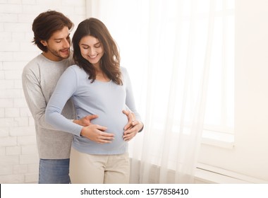 Loving man hugging his pregnant wife from behind standing near window at home, copy space - Powered by Shutterstock