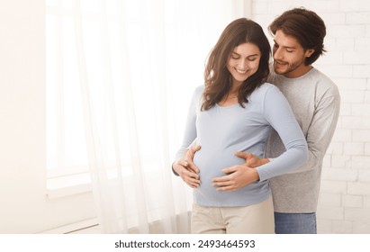 Loving man hugging his pregnant wife from behind standing near window at home, copy space - Powered by Shutterstock