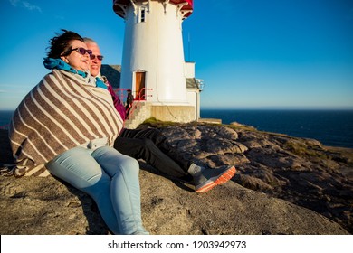 Loving mature couple hiking, sitting on windy top of rock with lighthouse. Mature man and woman wrapped in blanket, hugging and Happily smiling. Scenic view of sea, mountains. Norway, Lindesnes. - Powered by Shutterstock